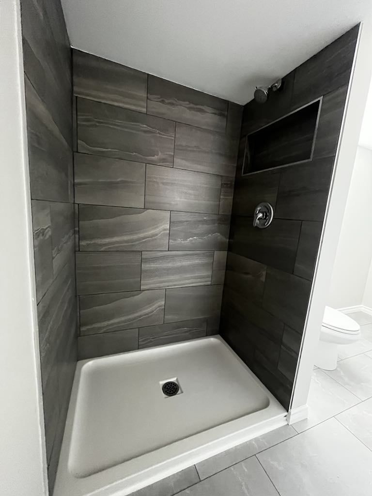 A compact shower stall with gray tiled walls, a built-in shelf niche, and a white shower base.