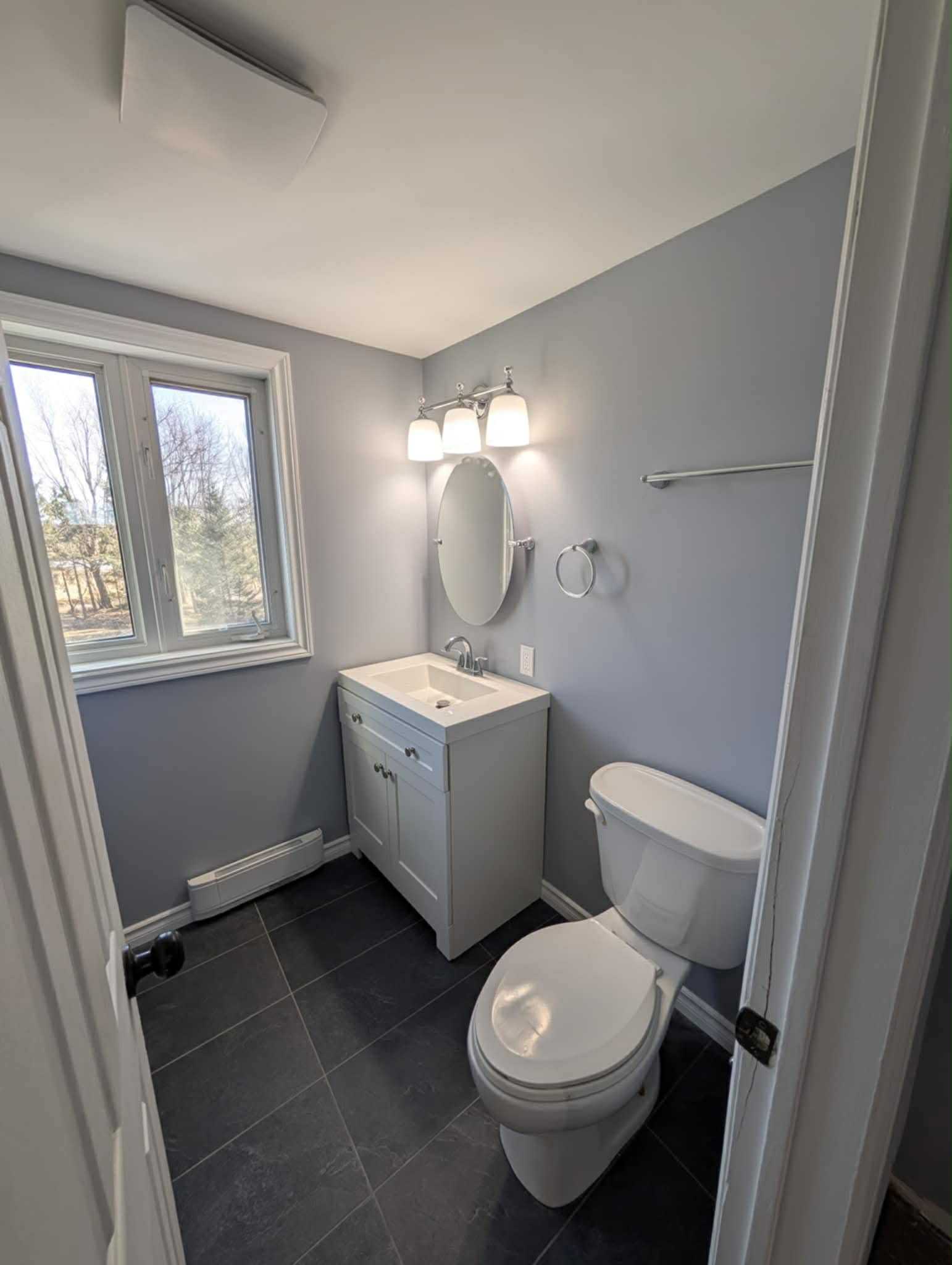 Updated bathroom remodel with white vanity, integrated sink, modern round mirror, new lighting, grey walls, and dark tile flooring completed by JSM Elite Contracting.