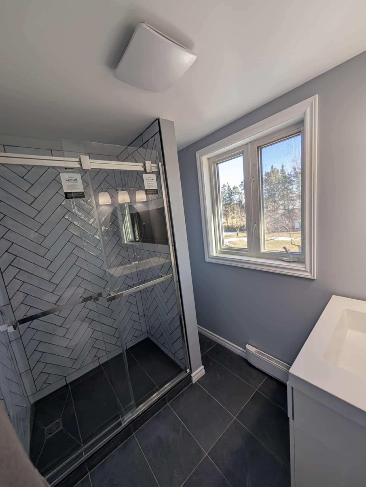 Modern bathroom remodeling project in Cornwall, Ontario featuring a glass shower with herringbone tile, dark floor tiles, soft grey walls, and a white vanity by JSM Elite Contracting.