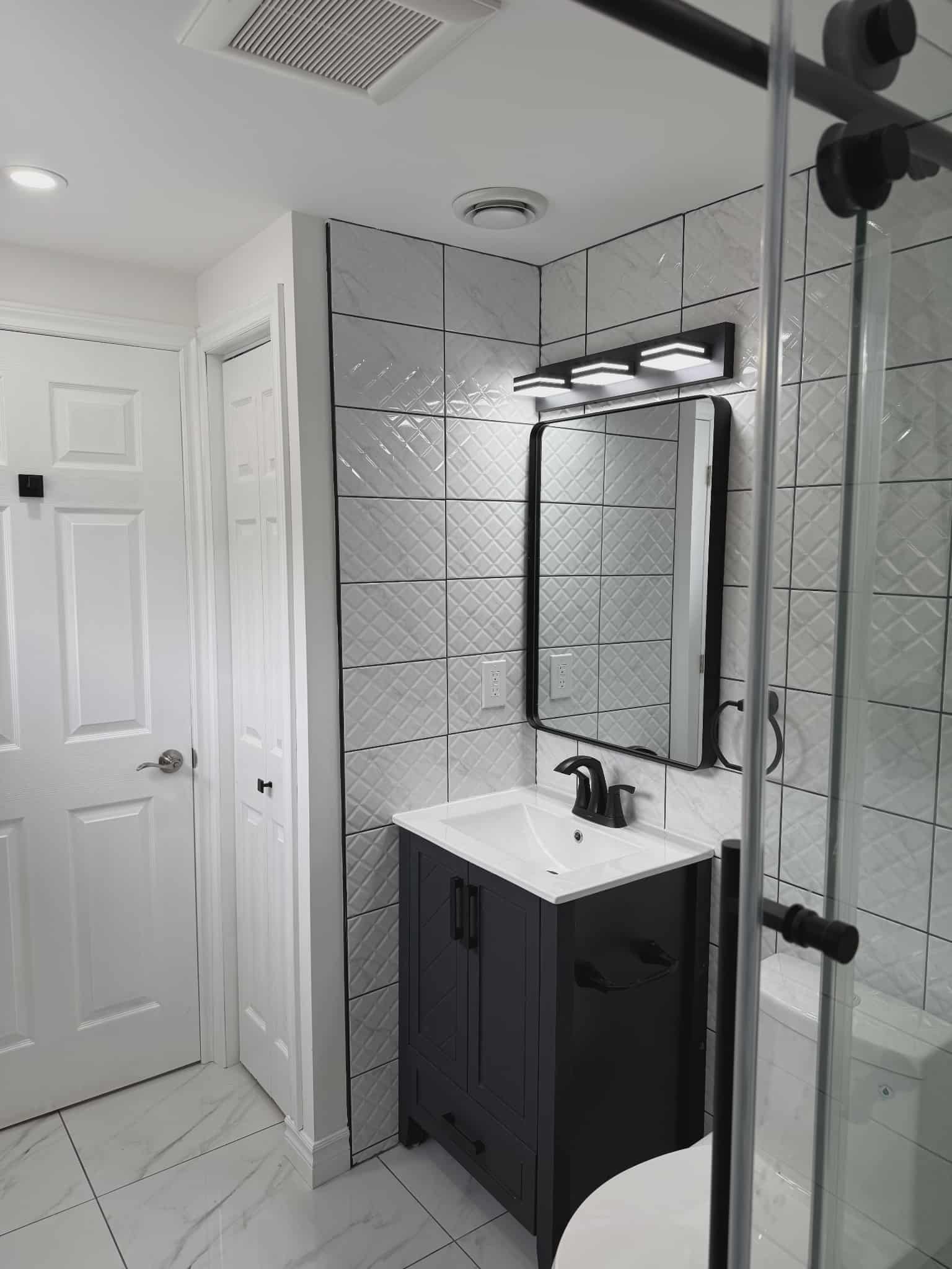 Contemporary bathroom with dark vanity, white countertop, black faucet and mirror frame, white geometric wall tiles, and marble-look floor, renovated by JSM Elite Contracting in Cornwall, Ontario.