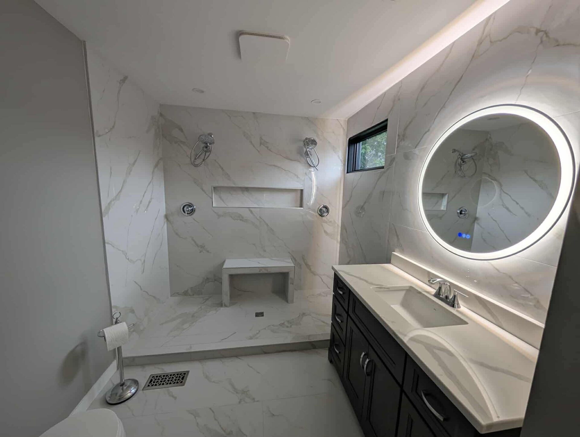 Modern Cornwall bathroom remodel featuring a walk-in shower with dual shower heads, marble-look tiles, a built-in bench, recessed shower niche, dark vanity with quartz countertop, and an LED backlit mirror—renovated by Elite Contracting Ltd.