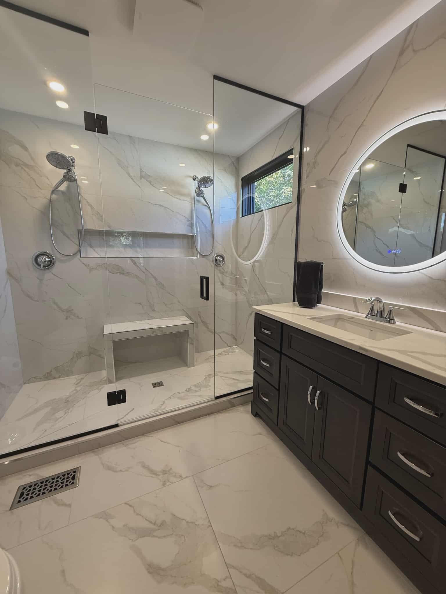 Luxury bathroom remodel featuring large-format marble tile, frameless glass shower with bench, dual shower heads, and modern vanity with backlit round mirror by JSM Elite Contracting.