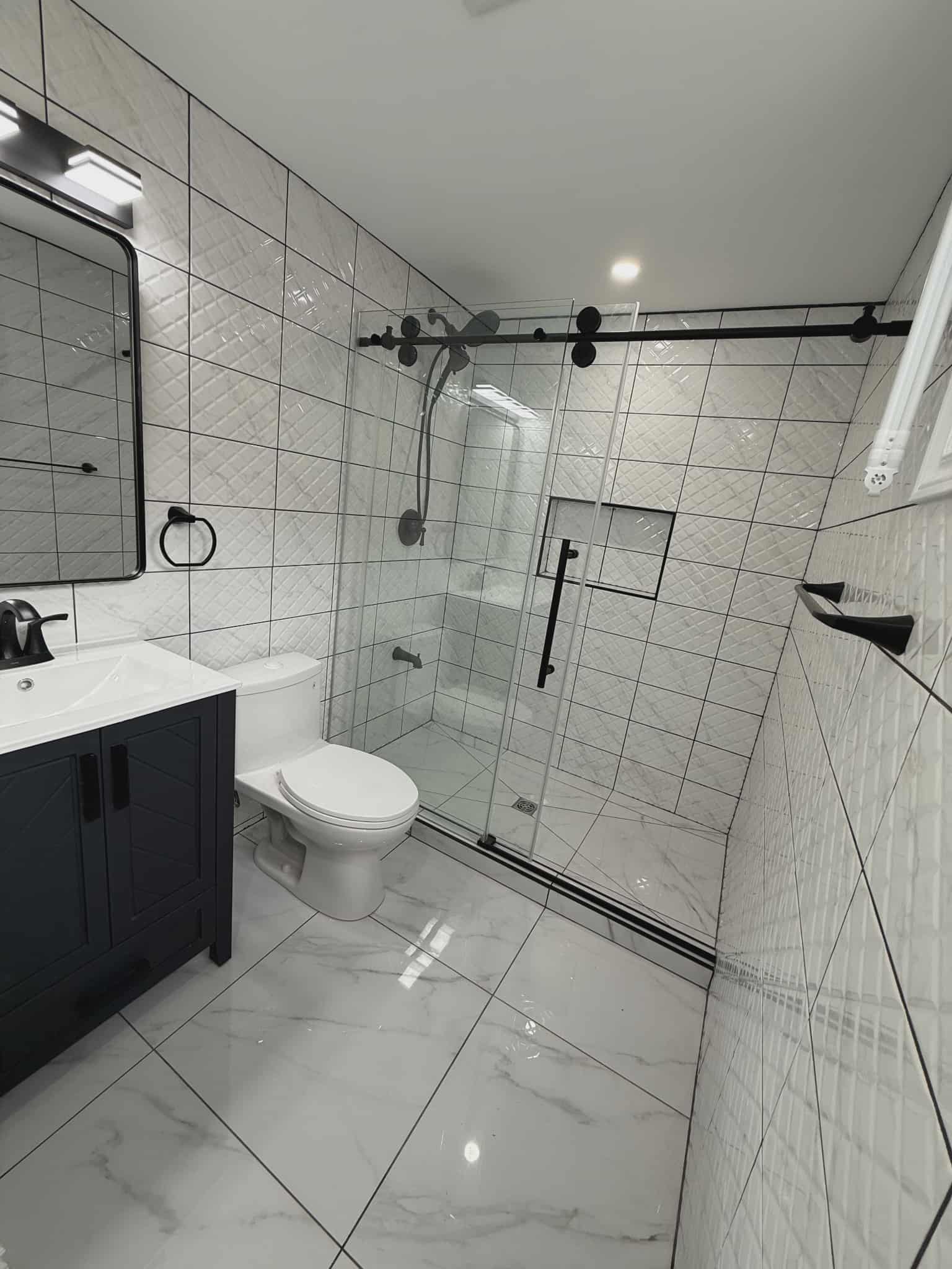 Modern bathroom with glossy marble-look floor tile, full-length glass shower enclosure with black sliding hardware, geometric white wall tiles, and dark vanity, renovated by JSM Elite Contracting in Cornwall, Ontario.