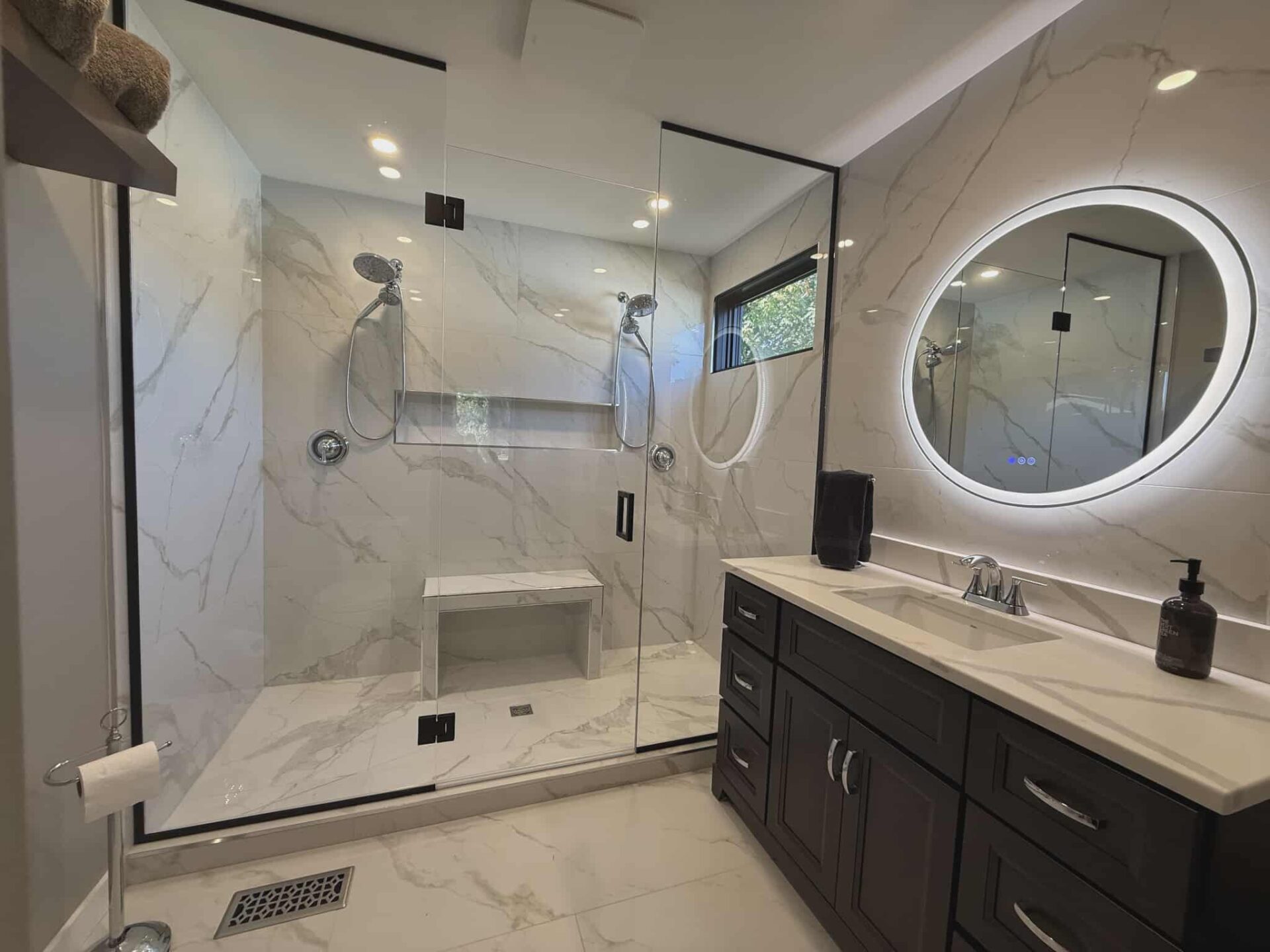 Modern bathroom renovation featuring a frameless glass walk-in shower with dual shower heads, built-in bench, marble-look tile, and dark wood vanity with backlit round mirror by JSM Elite Contracting.