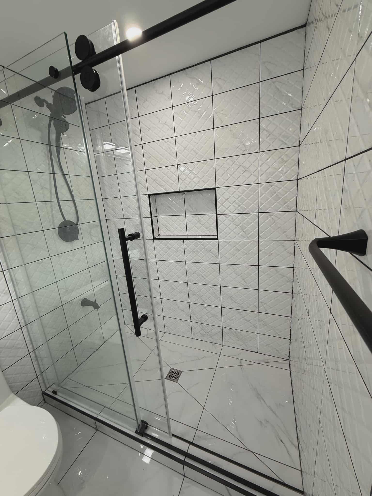 Sliding glass shower door with black hardware enclosing a custom tile shower with white geometric wall tiles, recessed niche, and marble-look floor, renovated by JSM Elite Contracting in Cornwall, Ontario.
