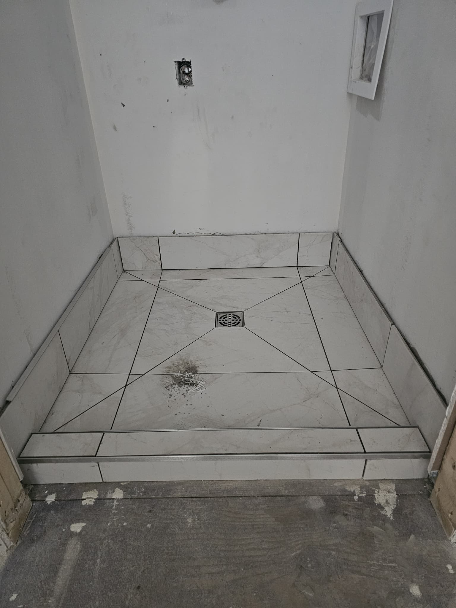 In-progress tiled shower base with centered square drain, marble-look floor tiles, and raised curb in a new walk-in shower installation by JSM Elite Contracting in Cornwall, Ontario.