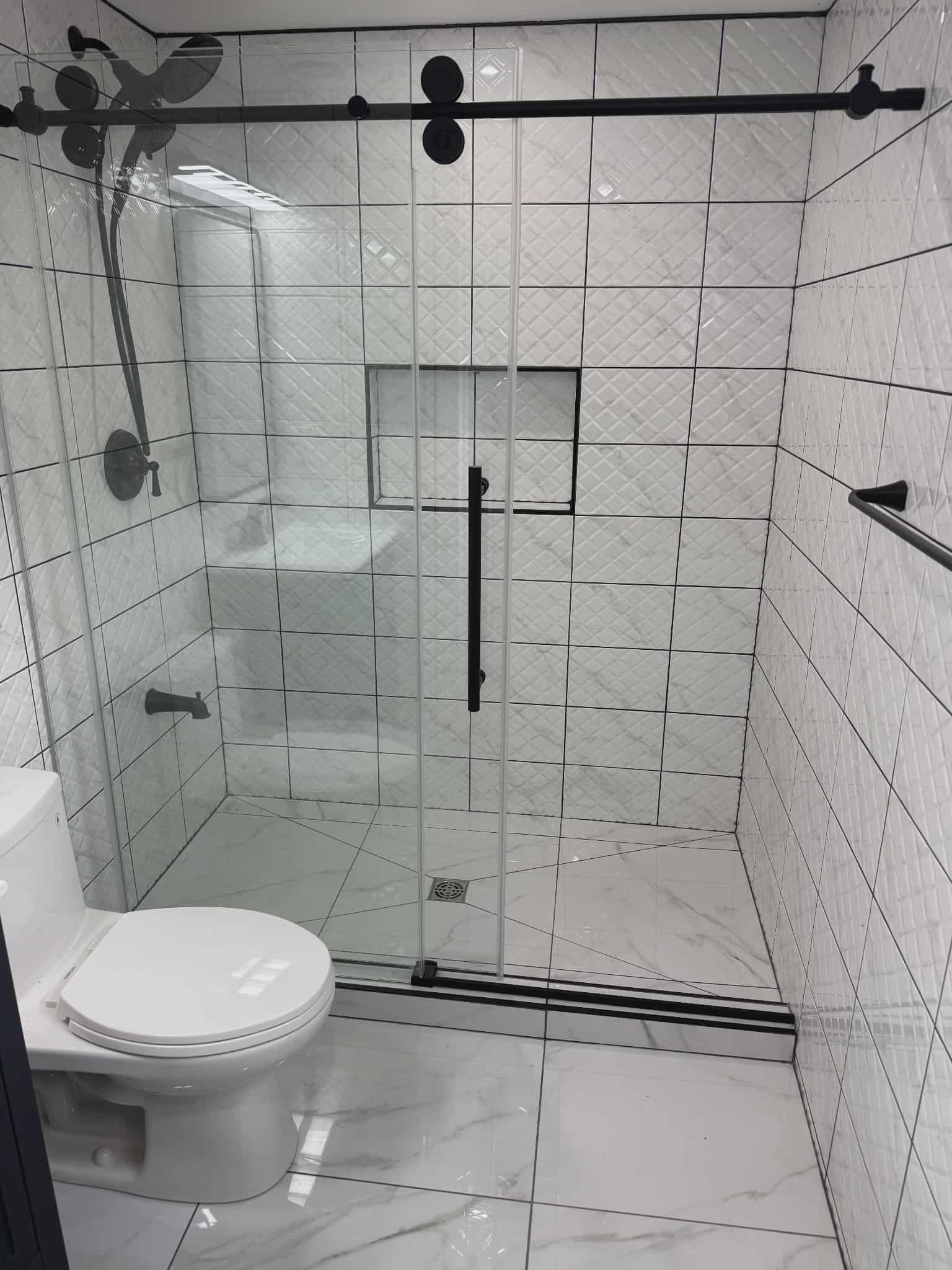 Modern bathroom with white geometric shower tiles, full-length glass enclosure with black sliding hardware, recessed niche, built-in bench, and marble-look floor, renovated by JSM Elite Contracting in Cornwall, Ontario.